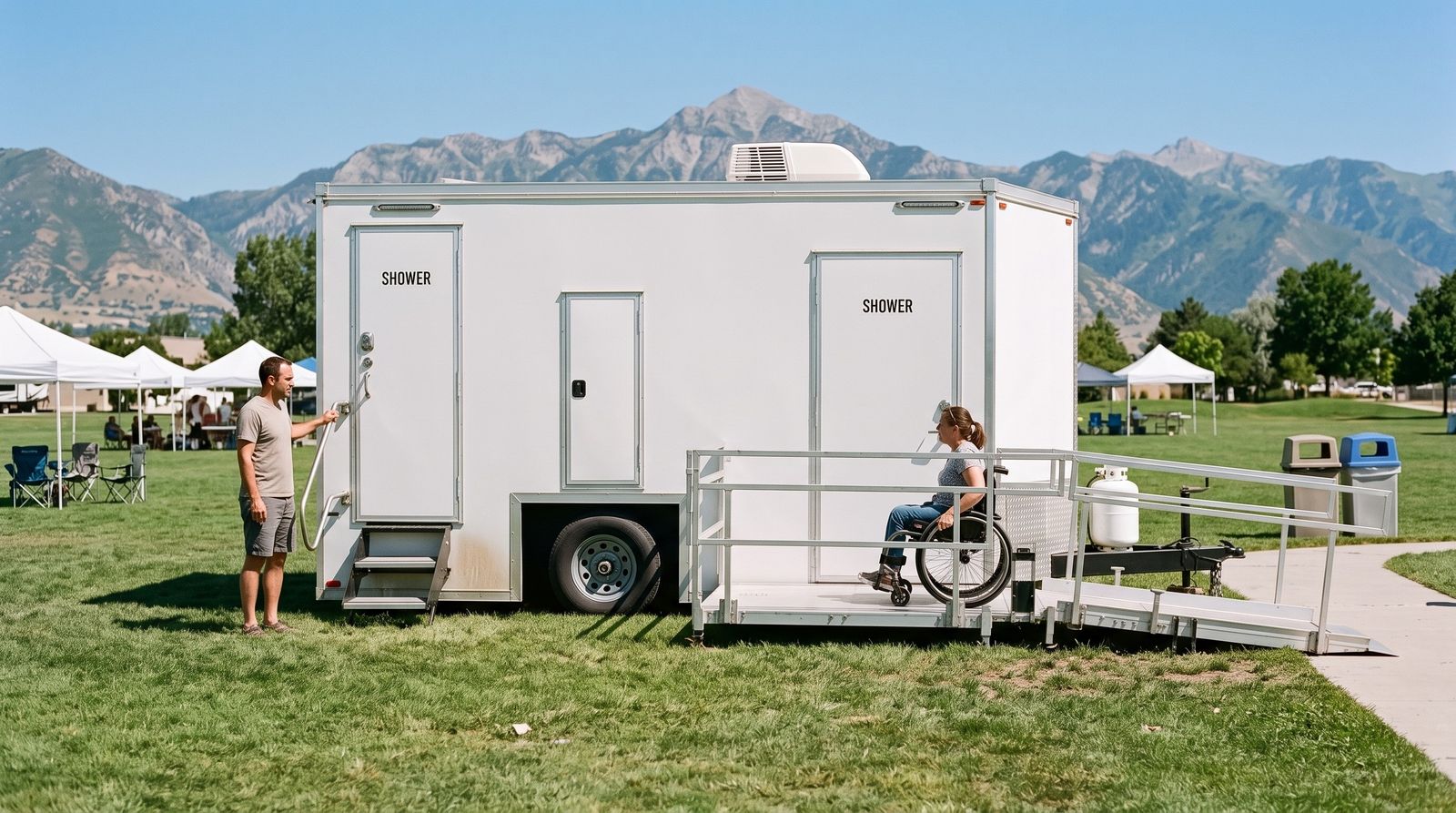 Accessible shower trailer at a Draper park festival, a wheelchair user on its platform and a man at the stepped door, Lone Peak behind
